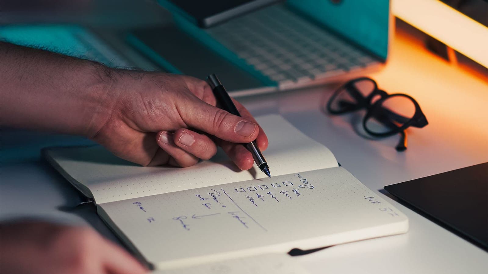 Hands write in an open notebook beside a phone and laptop.